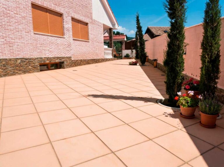 Terraza exterior pavimentada con baldosa porcelánica de gran formato en tonos beige claro, con acabado liso. La superficie muestra un patrón de rejuntado regular. Flanquean el espacio macetas de barro con flores rojas y cipreses, y se observan construcciones de ladrillo y hormigón al fondo bajo cielo despejado.