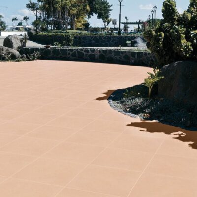 Terraza exterior pavimentada con baldosa de gran formato en tonos beige y arena, con acabado liso y continuo. Zona ajardinada con piedras decorativas, plantas y árboles al fondo, junto a pasarela de madera y farolas de jardín.