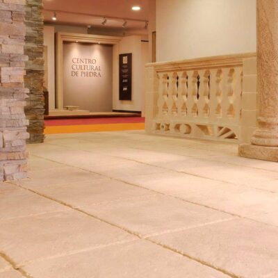 Espacio interior amplio con suelos de gres porcelánico en tonos beige y crema, columnas de piedra natural y revestimiento decorativo de piedra caliza en pared. Ambiente cultural con acabado mate y formato grande.