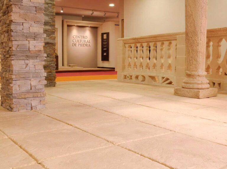 Espacio interior amplio con suelos de gres porcelánico en tonos beige y crema, columnas de piedra natural y revestimiento decorativo de piedra caliza en pared. Ambiente cultural con acabado mate y formato grande.