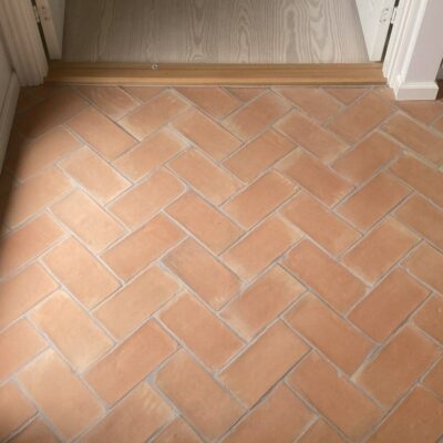Suelo interior revestido con baldosas de barro natural en tonos cálidos tierra, dispuestas en patrón de espiga. Las piezas rectangulares muestran la textura rústica característica del barro cocido, con ligeras variaciones de color y acabado mate natural.