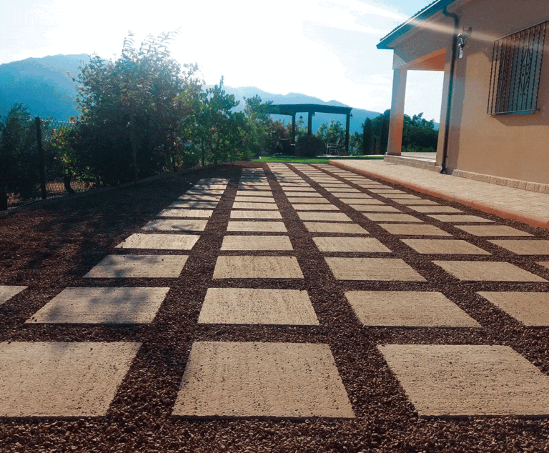 Terraza exterior pavimentada con baldosas de hormigón en tonos ámbar dispuestas en patrón geométrico, separadas por franjas de grava. Al fondo, pérgola y jardín con vistas a montañas.