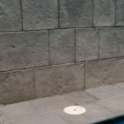 Aplacado de hormigón color antracita con textura de piedra en pared de piscina. Las baldosas de hormigón muestran acabado mate y formato rectangular con variación de tonos grises, instaladas en hiladas horizontales sobre el pavimento de hormigón.