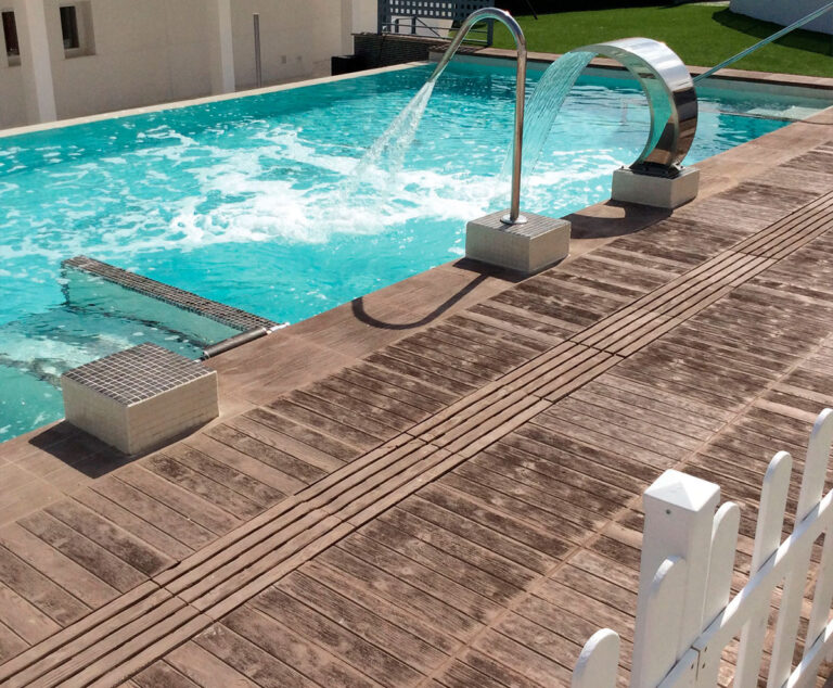 Terraza moderna con baldosas de hormigón imitación madera en tono beige junto a piscina de agua turquesa. Se aprecian fuentes decorativas de acero inoxidable sobre el pavimento exterior y zona ajardinada al fondo.