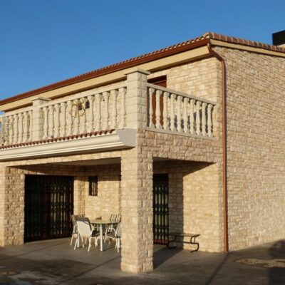 Vivienda unifamiliar de dos plantas con fachada revestida en aplacado imitación piedra natural en tonos beige y crema, balconada superior con balaustrada decorativa, cubierta de teja cerámica, y terraza inferior con zona de estar.