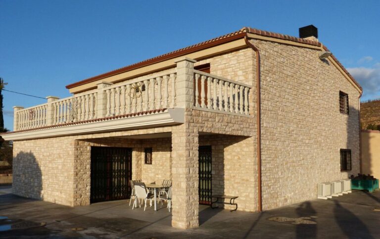 Vivienda unifamiliar de dos plantas con fachada revestida en aplacado imitación piedra natural en tonos beige y crema, balconada superior con balaustrada decorativa, cubierta de teja cerámica, y terraza inferior con zona de estar.