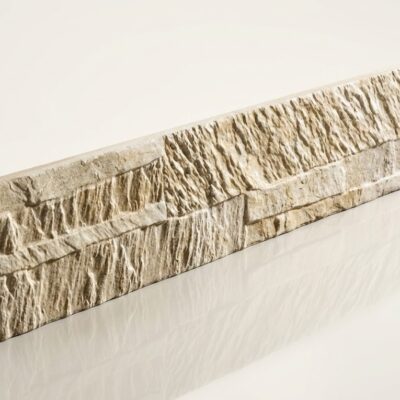Pieza rectangular de baldosa cerámica con acabado efecto piedra natural en tonos beige. Superficie texturizada con relieve irregular que imita la rugosidad de la piedra, presentando variaciones de color en tonos cálidos y grises. Formato alargado apto para revestimientos y suelos cerámicos.