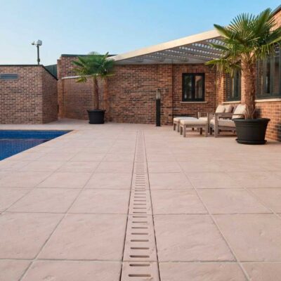Terraza exterior con pavimento de gres porcelánico color beige claro, drenaje lineal integrado, piscina azul, muros de ladrillo visto, sombrilla y mobiliario de exterior bajo pérgola.