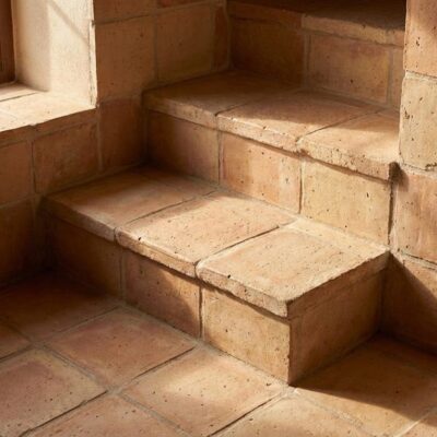 Escalera interior construida con baldosa de barro en tonos tierra, mostrando peldaños de formato rectangular y revestimiento de pared en bloques de terracota. Luz natural desde ventana lateral ilumina la textura rústica y acabado mate del material.