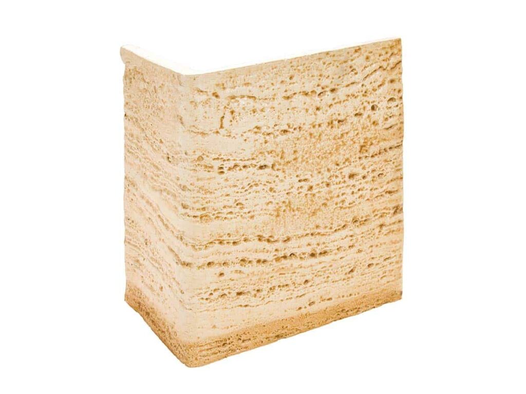 Pieza de esquina con acabado arenado en tonos beige y crema, imitando piedra travertino natural. Superficie texturizada con porosidad visible y aristas definidas para revestimiento de pared.