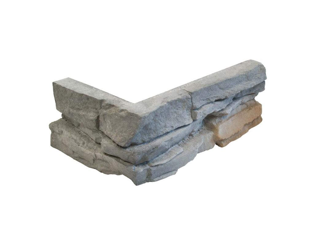 Pieza de revestimiento de hormigón con acabado imitación piedra natural en tonos grises. Formato rectangular con bordes irregulares y textura rugosa, simulando mampostería de piedra. Superficie mate con variaciones de color gris y beige.