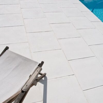 Terraza pavimentada con baldosas de suelo porcelánico de formato rectangular blanco, con textura lisa y acabado mate, adyacente a una piscina de agua azul turquesa.
