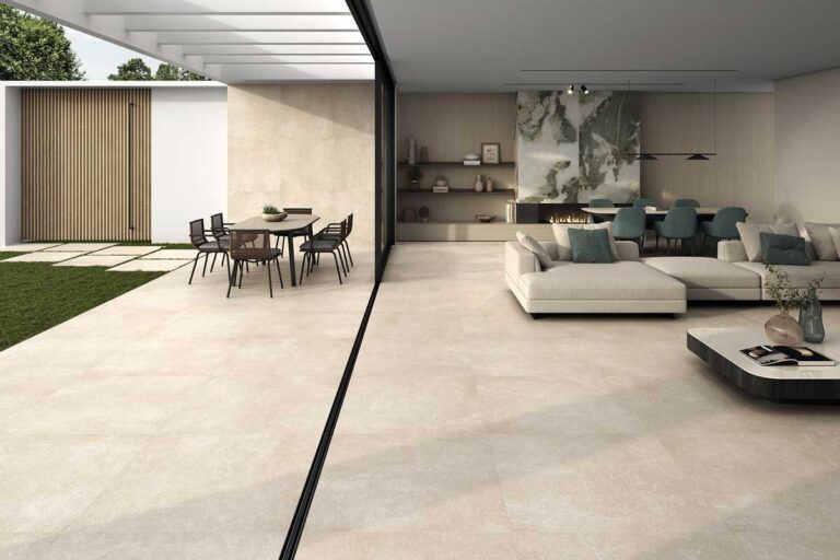 Espacio interior contemporáneo con suelos porcelanicos en tono beige claro que crea continuidad entre la zona de estar con sofá blanco y la terraza exterior. El acabado efecto piedra natural aporta calidez al ambiente minimalista.