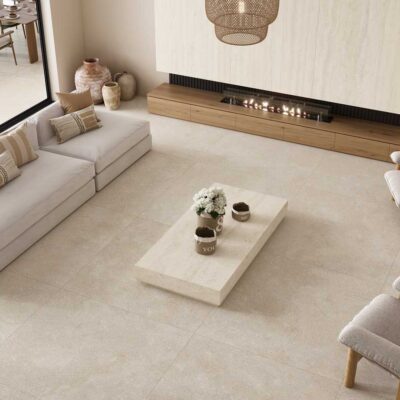 Salón contemporáneo con suelo de gres porcelánico imitación piedra en tonos ivory y beige claro. El espacio muestra sofás modulares en color crema, mesa de centro de piedra clara, sillones con estructura de madera y chimenea lineal integrada en el revestimiento de pared blanco.