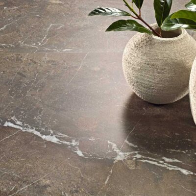 Baldosa rectangular de gres porcelánico con acabado mate imitando piedra natural en tonos grises y beige. Textura rugosa y superficie con veteado irregular característico del efecto piedra.