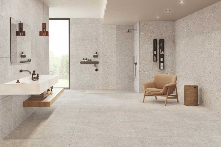 Baño contemporáneo con suelo de gres porcelánico efecto terrazo en tonos beige y grises, revestimiento de pared texturizado, ducha a italiena con mampara, mobiliario de madera clara y sillón de diseño.
