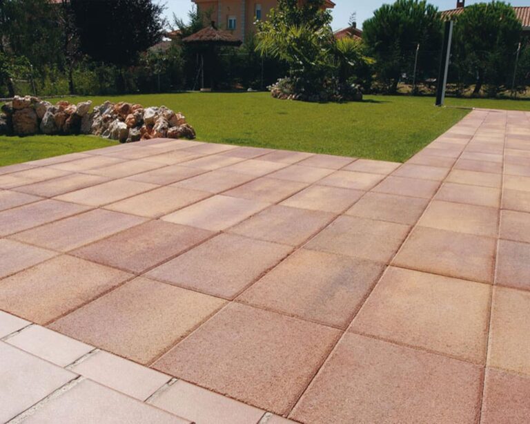 Terraza exterior pavimentada con losas de hormigón multicolor en tonos cálidos, dispuestas en patrón de espiga. Al fondo, jardín verde bien mantenido con vegetación y edificios residenciales.