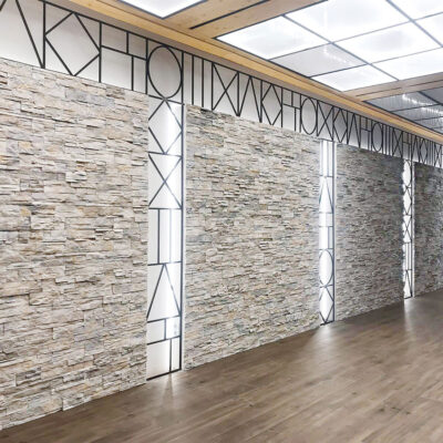Pasillo interior con paredes revestidas en piedra natural de tonos grises y beige, con vetas irregulares. Techo con lucernarios y estructura de madera, suelo de gres en tonalidad clara.