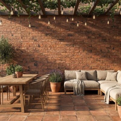 Terraza exterior con aplacado de pared efecto piedra en tonos cálidos y rojizos. La pared revestida muestra textura de piedra natural, complementada con suelo de barro cocido, mobiliario de madera y plantas en macetas de terracota.