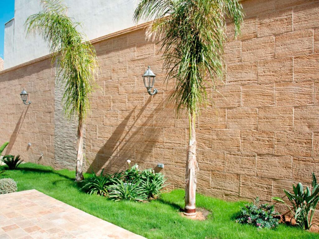 Fachada exterior revestida con aplacado que imita piedra travertino en tonos beige y arena, con textura arenada. La pared cuenta con iluminación mediante apliques metálicos y se complementa con vegetación tropical y césped en la base.
