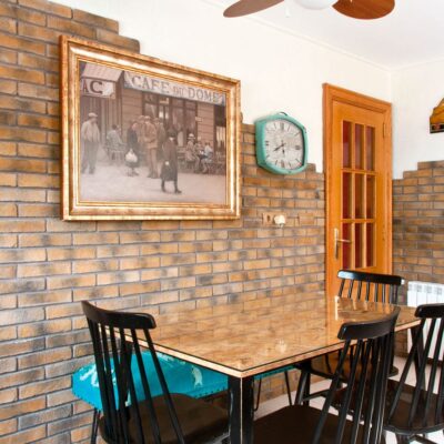 Comedor con pared de aplacado imitación ladrillo en tonos tierra y ocre, mobiliario negro de madera y suelo de gres en tonos cálidos. Reloj vintage y cuadro enmarcado completan la decoración.
