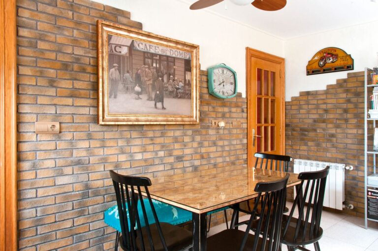 Comedor con pared de aplacado imitación ladrillo en tonos tierra y ocre, mobiliario negro de madera y suelo de gres en tonos cálidos. Reloj vintage y cuadro enmarcado completan la decoración.