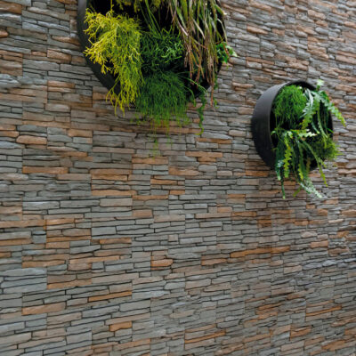 Pared interior revestida con aplacado de piedra en tonos gris marengo con acabado texturizado, decorada con dos maceteros circulares de plantas colgantes que generan contraste visual sobre la superficie de piedra.