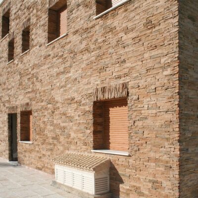 Fachada exterior con aplacado de piedra en tonos beige y ocre, mostrando ventanas rectangulares y una puerta con persiana de madera. El revestimiento de piedra presenta textura natural con acabado rústico.