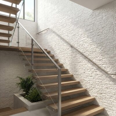 Escalera interior con peldaños de madera y revestimiento de pared efecto piedra crema en acabado texturizado. Barandilla metálica minimalista y ventanas superiores proporcionan iluminación natural al espacio.