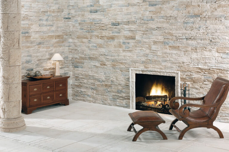 Salón contemporáneo con pared de revestimiento que imita piedra natural en tonos beige y gris, chimenea encendida, muebles de cuero marrón y lámpara de mesa sobre aparador de madera.