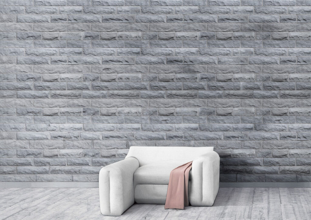 Espacio interior con pared revestida con azulejos efecto piedra gris ceniza en formato rectangular, suelo de gres claro y sillón blanco con manta rosa.