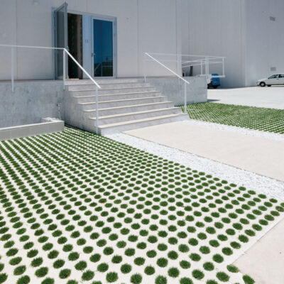 Área exterior con pavimento de baldosas de hormigón en patrón geométrico alternado verde y blanco, escaleras de acceso a edificio moderno de hormigón visto, y dos vehículos estacionados. Suelo porcelánico imitación con diseño decorativo y funcional para espacios públicos.
