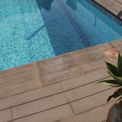 Terraza junto a piscina con peldaño de gres porcelánico imitación madera en tonos cálidos, suelo de mosaico azul turquesa y maceta con planta verde en primer plano.