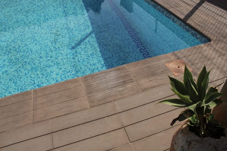 Terraza junto a piscina con peldaño de gres porcelánico imitación madera en tonos cálidos, suelo de mosaico azul turquesa y maceta con planta verde en primer plano.