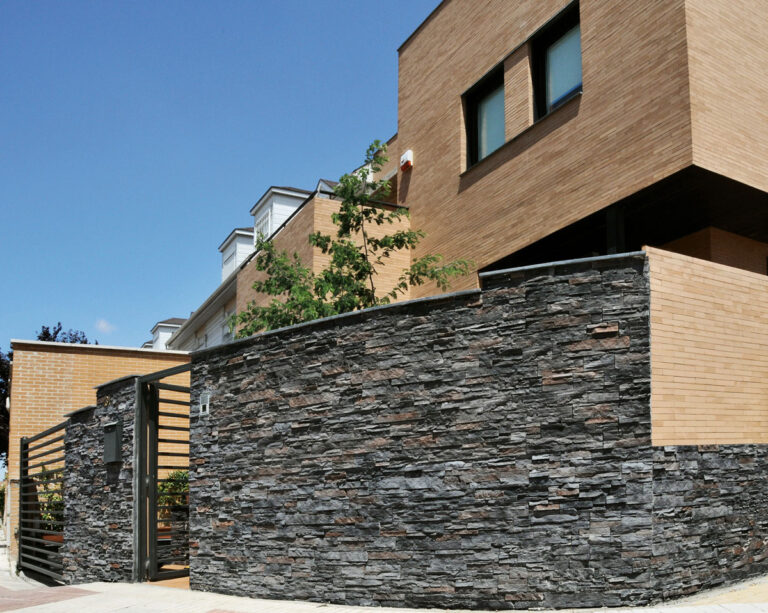 Fachada exterior contemporánea revestida con piedra natural color gris oscuro de textura irregular, combinada con ladrillo cerámico claro en las plantas superiores y cercado con aplacado de piedra en tonos oscuros.