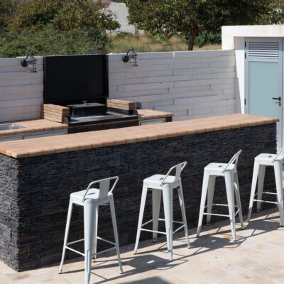 Terraza exterior con aplacado de piedra natural gris oscuro en muros y barra de cocina, cubierta de madera, equipada con grill integrado y cuatro taburetes metálicos blancos. Al fondo, ducha exterior de obra y vegetación natural.