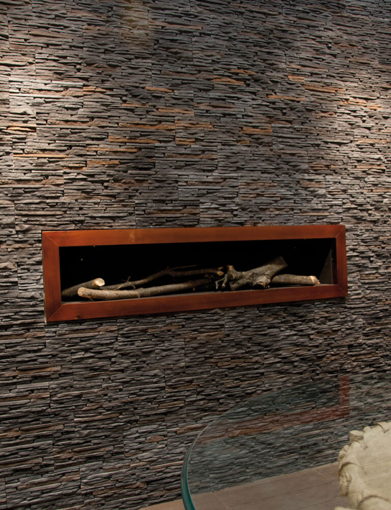 Revestimiento de pared con aplacado de piedra en tonos grises y marrones con textura irregular de stacked stone, instalado en ambiente interior con chimenea de leña integrada en marco metálico.