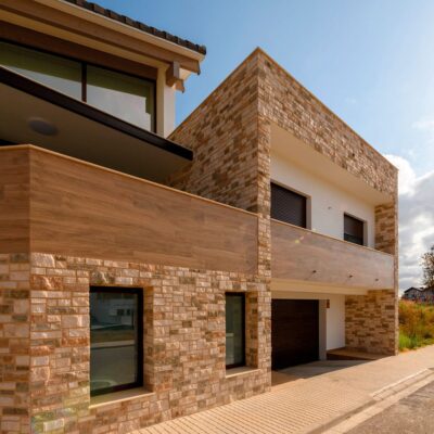 Fachada de vivienda unifamiliar revestida con placas de piedra natural Colorado corteza en tonos ocres y terracota. Combinación de aplacado de piedra en muros con franjas horizontales de madera clara y hormigón, bajo cielo despejado.