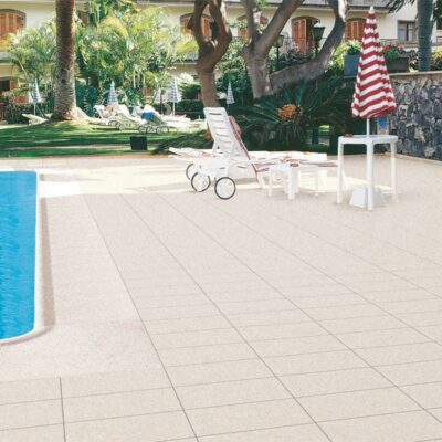 Terraza exterior con suelo porcelánico de gran formato en tonos beige claro, adyacente a piscina azul. Zona de descanso con sombrilla roja y blanca, edificio colonial y jardín con árboles de fondo.