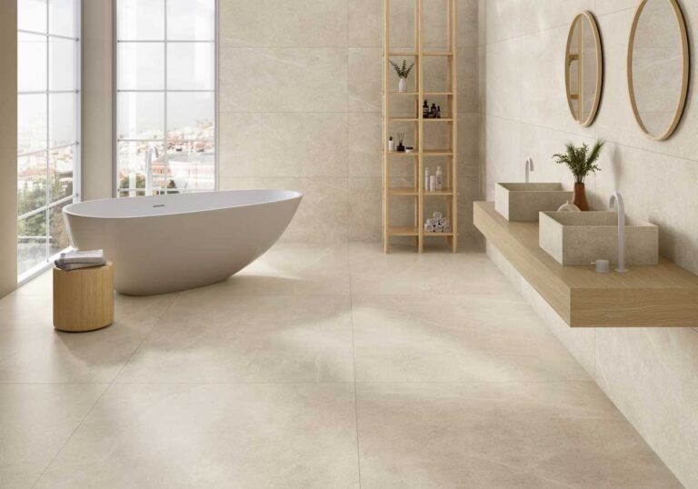 Baño contemporáneo con suelo y revestimiento en porcelánico efecto mármol Hope Ivory en tonos beige claros. Bañera blanca exenta junto a ventanal panorámico, lavabos de hormigón sobre encimera de madera, espejos circulares dorados y mueble de almacenamiento en madera natural.