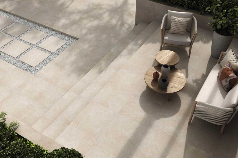 Terraza exterior con suelo de gres porcelánico acabado efecto piedra natural en tonos beige, mobiliario contemporáneo con sillas tapizadas grises y mesitas de madera redondas, vegetación en macetas de hormigón y sombras proyectadas del mobiliario.