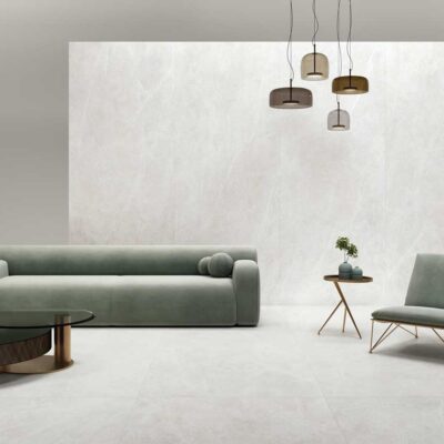 Espacio interior minimalista con suelo porcelánico gran formato efecto mármol blanco, sofá tapizado en verde pálido, silla metálica con respaldo tapizado, y luminarias colgantes en tonos neutros.