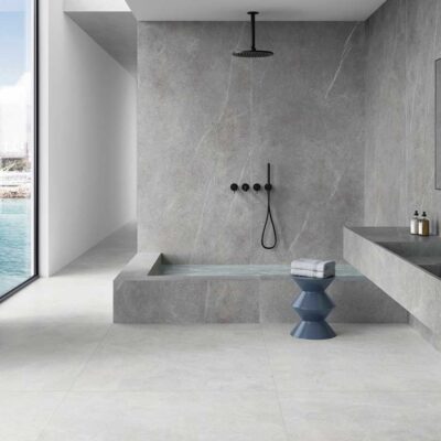 Baño contemporáneo con paredes y suelo en porcelánico imitación mármol gris claro en gran formato, ducha abierta con mampara, doble lavabo integrado y vistas al mar. El acabado mate del suelo cerámico crea una superficie continua y elegante.
