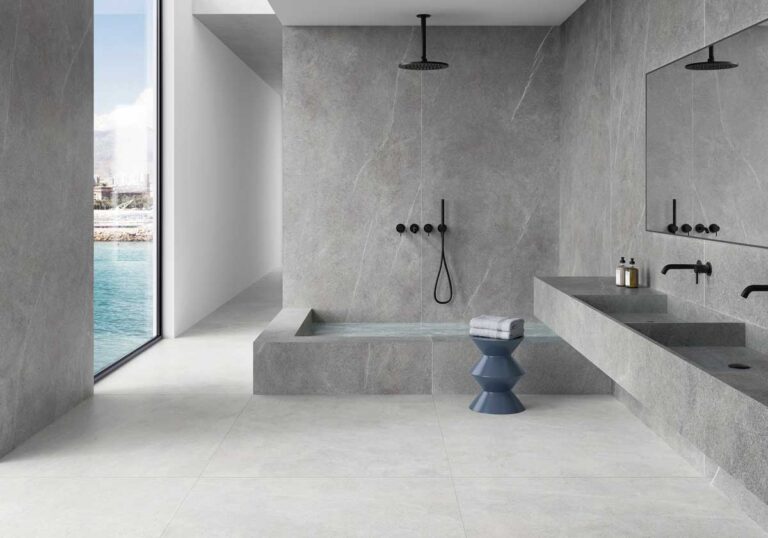 Baño contemporáneo con paredes y suelo en porcelánico imitación mármol gris claro en gran formato, ducha abierta con mampara, doble lavabo integrado y vistas al mar. El acabado mate del suelo cerámico crea una superficie continua y elegante.