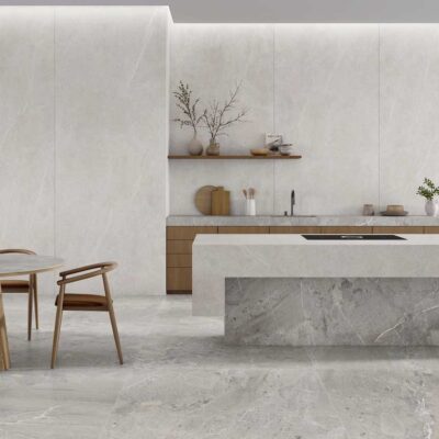 Cocina moderna con suelo de gres porcelánico efecto mármol blanco, isla central en tono gris claro, revestimiento de pared blanco mate y mesa redonda de madera con sillas de diseño. Zona de comedor integrada con plantas decorativas y grandes ventanales al fondo.