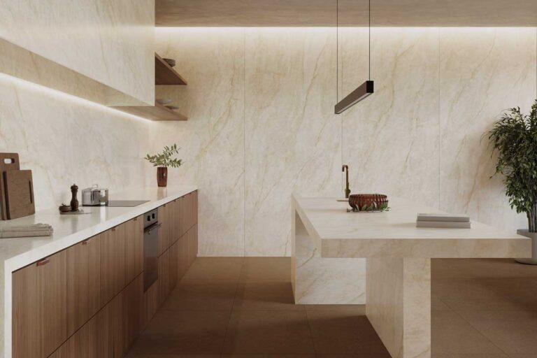 Cocina de diseño minimalista con paredes revestidas en gres porcelánico imitación mármol natural en tonos beige y crema. La encimera blanca contrasta con muebles de madera clara, y el suelo continúa con el mismo acabado porcelánico en formato rectificado.
