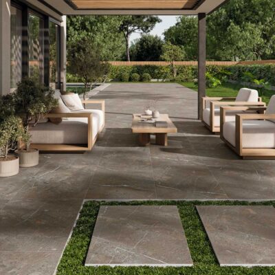 Terraza cubierta con porcelánico imitación piedra en tonos grises y naturales, mobiliario de madera clara con cojines beige, vistas al jardín verde, acabado mate y formato grande.