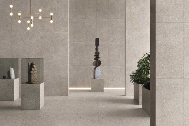 Espacio de galería moderna con suelo de gres porcelánico en tonos beige y terrazo, grandes placas rectificadas en formato continuo. Paredes de revestimiento cerámico liso gris claro, iluminación colgante dorada, pedestales de hormigón con esculturas y plantas decorativas.