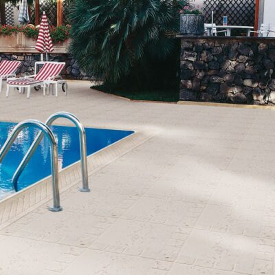 Zona de piscina exterior con remate cerámico de color beige alrededor del borde del agua, tumbonas a rayas rojas y blancas al fondo, y vegetación tropical. El suelo de piscina presenta baldosas cerámicas en tonos claros con acabado antideslizante.