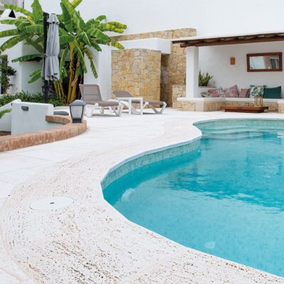 Terraza de exterior con piscina curva de agua turquesa rodeada de pavimento de hormigón en tonos claros, aplacados de piedra natural en muros, plantas verdes y zona de descanso con cojines de colores.